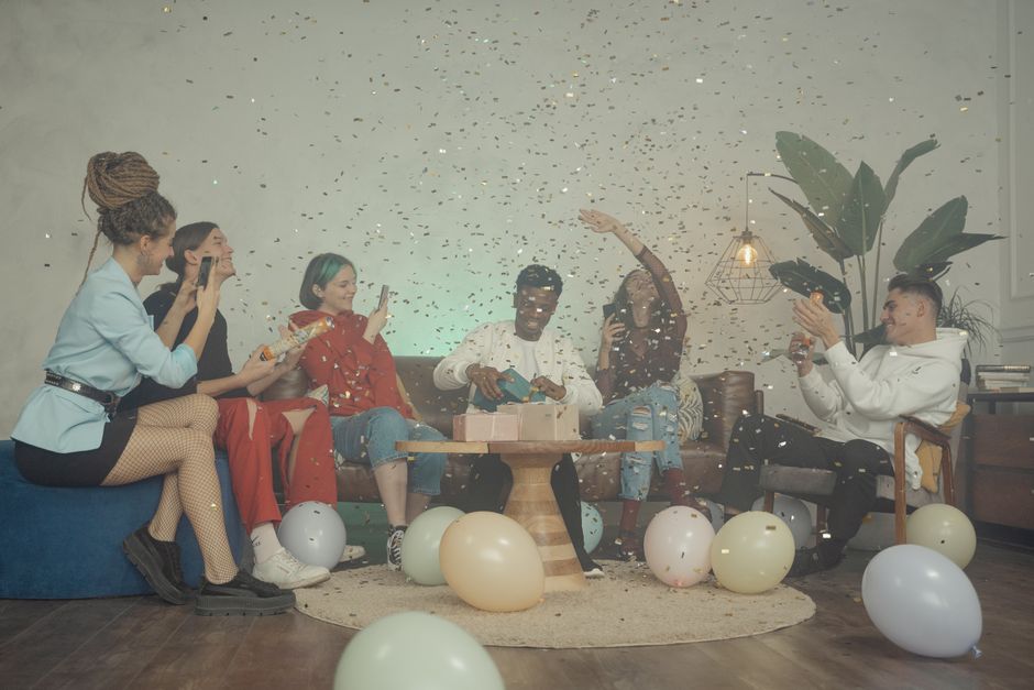 Group of friends celebrating a birthday indoors with balloons and confetti.